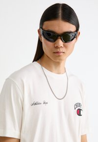 Schwarze Sonnenbrille mit modernem Design, ein weißes T-Shirt mit dem Aufdruck "Athletics Dept." und einem kleinen Logo sowie eine silberne Kettenhalskette.