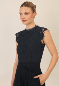 Vestido de encaje azul marino con escote en forma de concha y mangas cortas. Parte inferior de tela suave con bolsillos laterales y diseño plisado. Silueta elegante y ajustada.