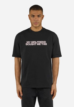 Mann trägt ein schwarzes T-Shirt mit hellrosa Schrift "MEINE FREUNDIN HAT MIR DAS GEKAUFT" und steht vor einem schlichten weißen Hintergrund.