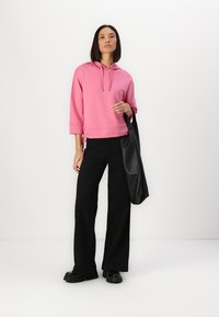 Felpa rosa con cappuccio, dal taglio comodo, maniche a tre quarti e lacci laterali, abbinata a pantaloni neri a gamba larga e una borsa nera.