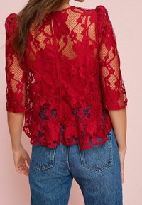Blouse en dentelle rouge avec motifs floraux, manches trois-quarts et ourlet à basque, associée à un jean bleu taille haute.