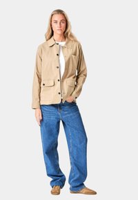 Beige canvasjakke med knaplukninger, to forlommer, parret med løstsiddende blå jeans og brune fodtøj.