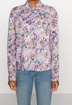 Mujer con camisa de botones lila con un patrón floral multicolor y vaqueros azules, de pie contra un fondo liso.