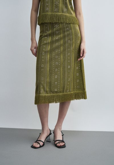 Olive green knitted skirt with floral patterns and fringe trim worn with black strappy block-heel sandals on a grey floor.