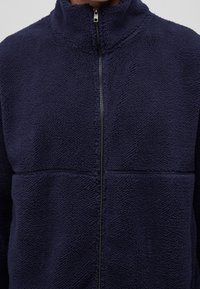 Giacca zip-up in pile blu navy con colletto alto, caratterizzata da un tessuto texture e una chiusura centrale con zip. Non sono visibili tasche.