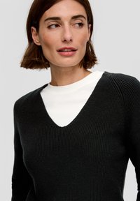Schwarzer Strickpullover mit tiefem V-Ausschnitt, strukturiertem Stoff und leicht puffierten Ärmeln, darüber getragen über einem weißen Rundhalsausschnitt-Shirt.
