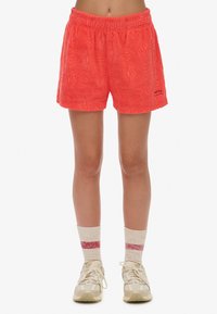 Shorts en corail, en tissu pelucheux, avec un motif de vagues texturé, dotés d'une taille élastique. Associés à des baskets crème et des chaussettes rayées.