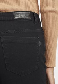 Jeans noirs avec un patch en cuir, tissu texturé et poches arrière classiques. La ceinture présente un détail de design subtil.