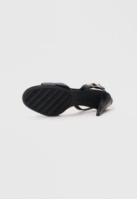 Svart høyhælt sandal med teksturert såle, ankelrem og gullspenne, plassert såle opp på en hvit bakgrunn.