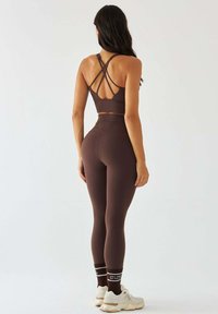 Brązowy sportowy crop top z krzyżującymi się na plecach ramiączkami, zestawiony z wysokim stanem legginsów. Modelka ma na nogach białe trampki i skarpetki w paski.