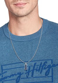 Sølv kæde med en rektangulær medaljon med en mørkeblå indsats, båret over en struktureret blå sweater.