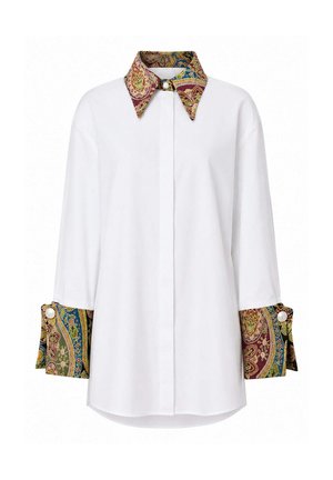 Femme portant un chemisier blanc à manches longues avec un col et des poignets paisley colorés, un jean large bleu foncé et des talons ouverts dorés, debout.