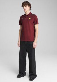 Burgundská polokošeľa, krátke rukávy, rebrovaný golier, s žltým logom Ferrari, skombinovaná s voľnými čiernymi nohavicami. Hladká textúra látky.