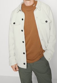 Vit fleecejacka med svarta knappar, bärs över en brun crewneck-tröja. Modellen har också på sig gröna cargobyxor.