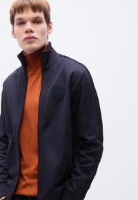 Σκούρο ναυτικό τζάκετ με φερμουάρ και ψηλό γιακά, με ανάγλυφο λογότυπο στο στήθος, σε συνδυασμό με ένα πορτοκαλί ζιβάγκο από μέσα.