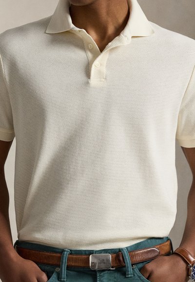 Lys kremfarget pikéskjorte med teksturert stoff, tre-knappers stolpe og korte ermer. Kombinert med et brunt belte og blågrønne bukser.