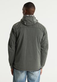 Chaqueta con capucha ligera de color verde oliva y corte relajado, con costuras visibles y un tejido texturizado, diseñada para el uso cotidiano.