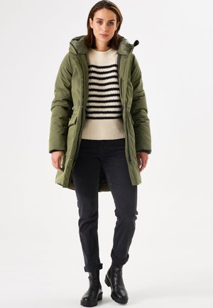 Doudoune vert olive avec capuche, portée sur un pull en tricot crème et noir à rayures, accompagnée de jeans foncés et de bottines noires.