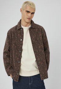 Veste à motif marron avec un devant à boutons, un design avec col et des manches longues, superposée sur un haut clair et un jean en denim foncé.