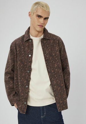 Veste à motif marron avec un devant à boutons, un design avec col et des manches longues, superposée sur un haut clair et un jean en denim foncé.