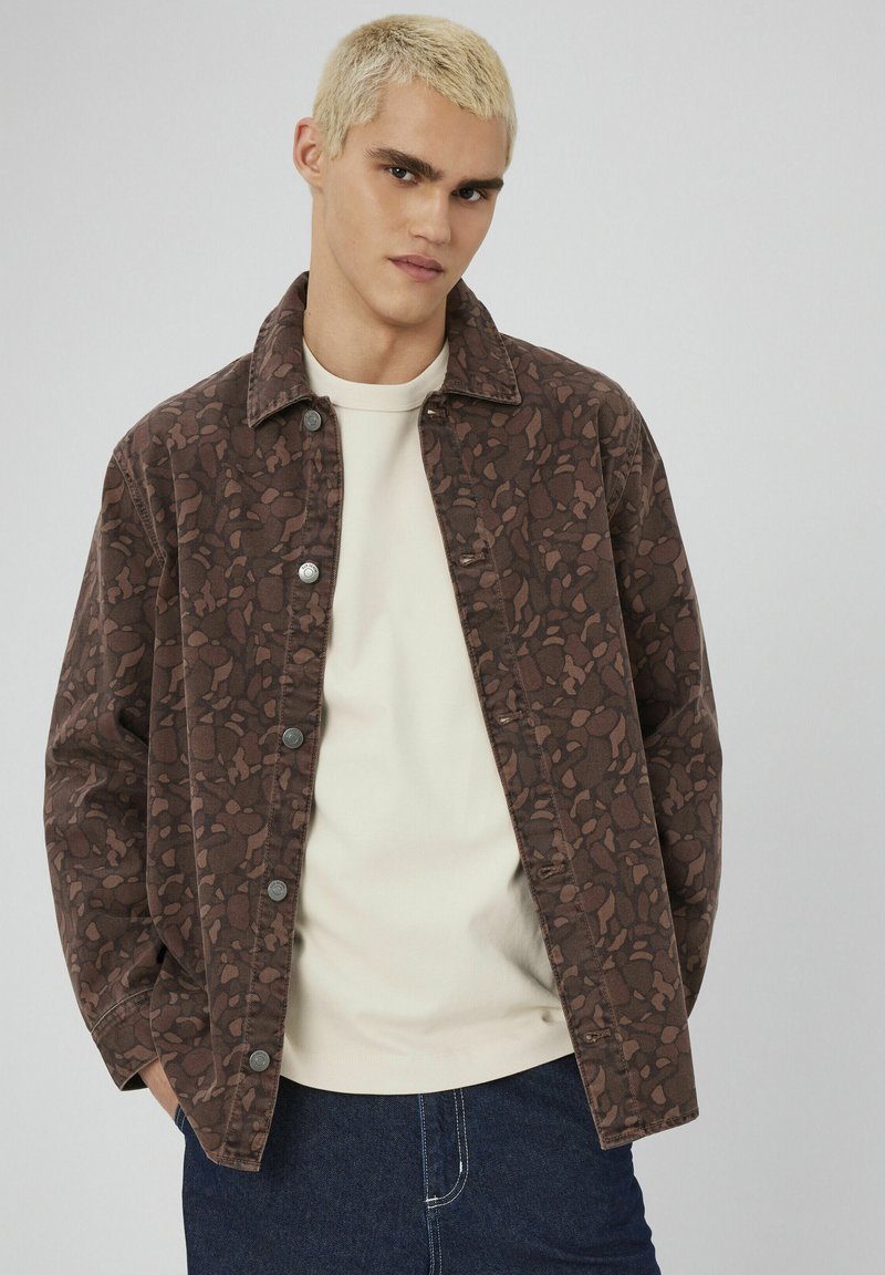 Veste à motif marron avec un devant à boutons, un design avec col et des manches longues, superposée sur un haut clair et un jean en denim foncé.