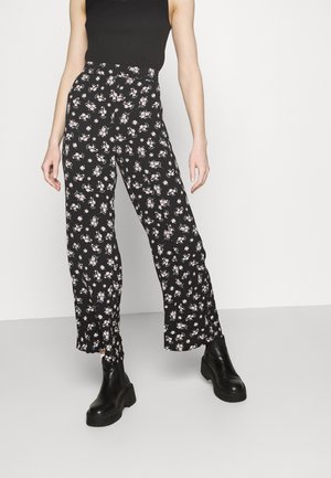 Person iført sorte bukser med høj talje med små hvide og lyserøde blomsterprint, sort ærmeløs top og sorte platform ankelsko.