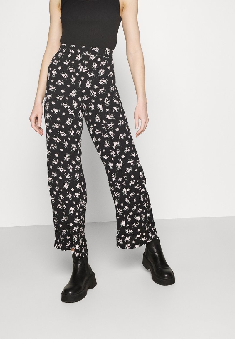 Personne portant un pantalon noir taille haute avec un petit imprimé floral blanc et rose, un top noir sans manches et des bottines à plateforme noires.