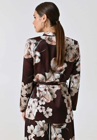 Giacca lunga a maniche lunghe floreale in marrone scuro con motivi floreali crema e rosa, caratterizzata da una vita con cintura e una texture liscia e satinata.