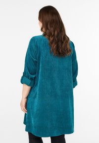 Zielona sukienka tunikowa z weluru z podwiniętymi rękawami, długości do połowy uda, o miękkiej teksturze i zaokrąglonym dołem. Prosty, casualowy design z minimalnymi akcentami.