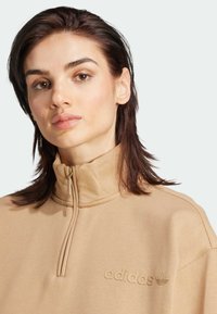 Felpa zip-up beige con colletto alto, tessuto testurizzato e dettaglio logo in rilievo sul petto. Design moderno e sportivo.