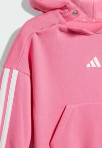 Sudadera rosa con forro de felpa, con acentos de tres franjas blancas en las mangas y un bolsillo tipo canguro. Incluye capucha con cierre a presión.