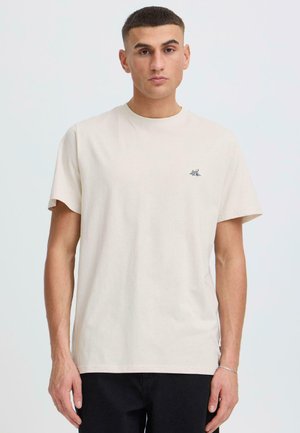 Nuori mies, jolla on lyhyet tummat hiukset, pukeutuneena yksiväriseen beigeen t-paitaan, jossa on pieni vuoristologo, seisomassa valkoista taustaa vasten.