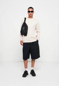 Sudadera beige clara, pantalones cortos negros y zapatillas negras. Bandolera negra llevada sobre el hombro, con gafas de sol. Estilo simple y atlético.