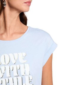 T-shirt bleu clair avec des manches retroussées, arborant un texte doré métallisé "AMOUR AVEC STYLE", en tissu lisse et à coupe décontractée.