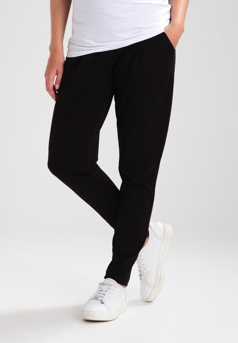 Černé joggingové kalhoty vyrobené z měkkého materiálu, s volným střihem, postranními kapsami a zúženým střihem nohavic, v kombinaci s bílými teniskami.