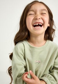 Jasnozielony, miękki sweatshirt z haftowanym wzorem kwiatu. Posiada okrągły dekolt i długie rękawy ze zgromadzonymi mankietami.