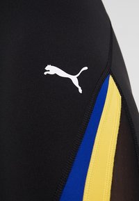 Svart tyg med vit Puma-logotyp och vertikala blå och gula ränder separerade av svart sömnad på sportkläder.