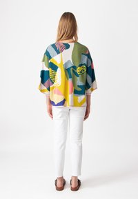 Kleurrijke, oversized gebreide top met abstracte vormen in groen, geel, roze en blauw. Draagt met witte broeken en sandalen.