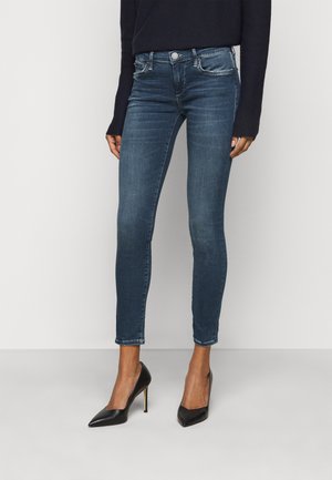 Kobieta ubrana w ciemnoniebieskie obcisłe dżinsy, czarną bluzkę z długim rękawem oraz czarne buty na wysokim obcasie i spiczastym noskiem, stojąca na jednolitym tle.