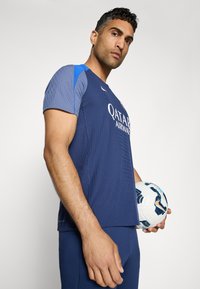 Mørkeblå sports T-shirt med struktureret stof, blå detaljer på skuldrene og "QATAR AIRWAYS" logo. Holder en mønstret fodbold.
