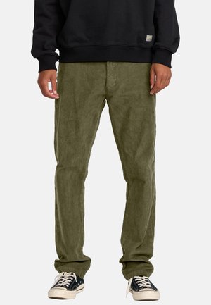 Olivgrüne Cordhosen mit einem lockeren Schnitt und gerippter Textur, kombiniert mit einem schwarzen Sweatshirt und schwarzen Sneakers mit weißen Sohlen.