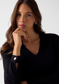 Schwarze Bluse mit V-Ausschnitt, langen Puffärmeln und einem markanten Knopfdetail an der Manschette. Das Modell hat welliges braunes Haar und posiert nachdenklich.