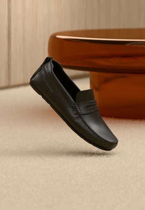 Zapato mocasín de cuero negro flotando sobre una alfombra beige con una pieza de mobiliario curva color ámbar marrón en el fondo.