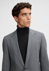 Grau strukturierter Blazer mit gekerbtem Revers, Ein-Knopf-Verschluss, getragen über einem schwarzen Rollkragenpullover. Glatter Stoff, taillierte Passform.