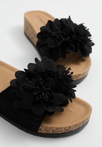 Sandalias de cuña negras con grandes adornos de flor negra, parte superior de ante, media suela de corcho y plantillas en tono marrón. La suela plana proporciona estabilidad.