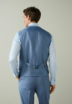 Férfi világoskék ingben, kék mellényben áll, amelyen állítható hátpánt látható, és hozzáillő kék nadrágot visel, háttal egy egyszerű háttér előtt.