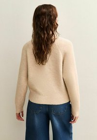 Suéter de punto beige con una textura suave, dobladillo acanalado y mangas largas. Combinado con jeans de pierna ancha de color azul oscuro, que muestran un ajuste relajado.