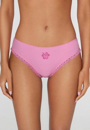 Kobieta w różowej bieliźnie bikini z koronkową lamówką i haftowanym kwiatem z przodu, stojąca na jasnym tle.