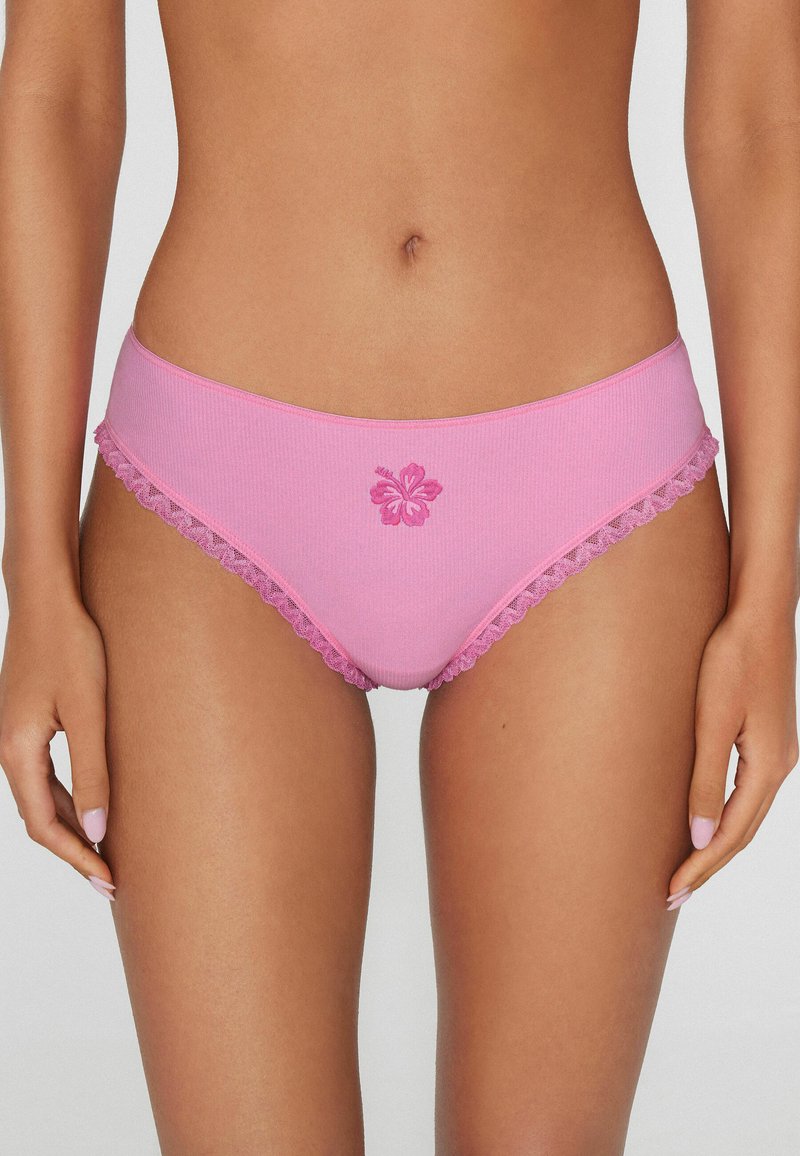 Vrouw die roze bikini-ondergoed draagt met kanten randje en geborduurde bloem aan de voorkant, staand tegen een lichte achtergrond.