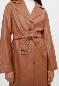 Hnědý kabát z umělé kůže s páskem v pase, širokými klopami a knoflíkovým zapínáním. Má hladkou strukturu a kontrastní sponu.